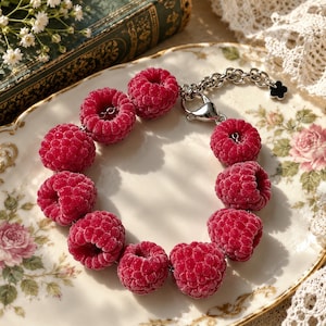 May include: A bracelet featuring a series of realistic, textured red raspberry charms linked together. The piece includes a silver-toned metal chain with a lobster claw clasp and a small, black, four-petaled flower charm attached to the end of the chain.
