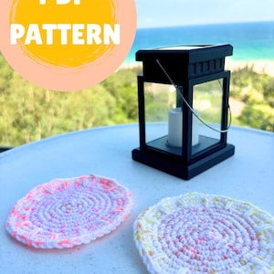 May include: A black lantern with a white candle sits on a white surface next to two crocheted coasters. One coaster is pink and white with neon orange accents, and the other is white and pink with yellow accents. The image includes a yellow circle with the words "PDF PATTERN".