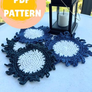 May include: A collection of four handmade crochet coasters in shades of blue and white. The coasters have a circular design with a white center and a fringed edge. A black lantern with a lit candle sits in the background. The image includes the text "PDF PATTERN".