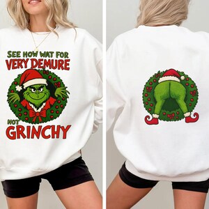 Può includere: Felpa bianca con un design a tema Grinch. La parte anteriore presenta il Grinch con un cappello da Babbo Natale all'interno di una ghirlanda, con il testo "SEE HOW WAT FOR VERY DEMURE NOT GRINCHY". Il retro mostra il sedere del Grinch con una ghirlanda e un cappello da Babbo Natale.