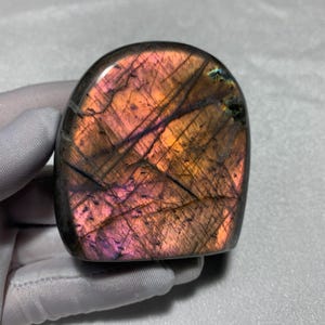 May include: A polished labradorite stone with a smooth, rounded top and a flat base. The surface displays a vibrant iridescence featuring shifting hues of warm orange, deep pink, and purple, intersected by dark, thin, natural mineral inclusions.
