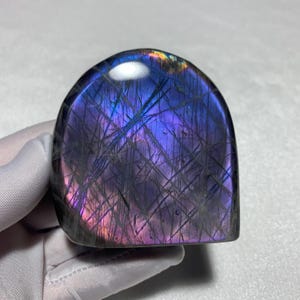 May include: A polished labradorite stone with a smooth, dome-shaped top and a flat base. The surface displays a vibrant iridescent sheen with deep blue and purple hues, accented by a network of dark, intersecting linear inclusions throughout.