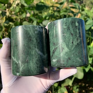 May include: Two cylindrical cups crafted from polished dark green stone featuring natural white and black veining patterns throughout the smooth surface. The vessels have a straight-sided design with a uniform diameter and a glossy finish.