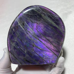 May include: A polished, teardrop-shaped labradorite stone featuring a vibrant, iridescent purple flash across its surface. The stone displays deep violet hues with dark, intersecting mineral veins and a smooth, reflective finish throughout.