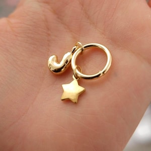 May include: A gold-toned hoop earring featuring two dangling charms. One charm is a smooth, rounded letter J, and the other charm is a five-pointed star. The metal has a polished, reflective finish, and the hoop is circular with a simple clasp mechanism.