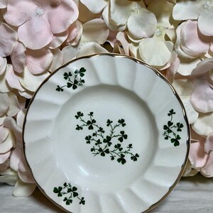 May include: A white ceramic saucer with a gold rim and a green shamrock design. The saucer has a scalloped edge.