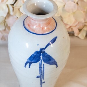 Può includere: Un vaso in ceramica bianca con un motivo floreale blu e bianco. Il vaso ha un corpo arrotondato e un collo stretto. La parte superiore del vaso ha un bordo rosa chiaro.