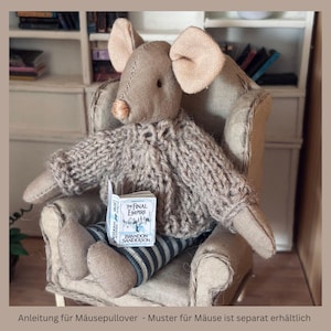 May include: A beige fabric mouse doll wearing a chunky knit sweater and blue and white striped pants. The mouse is seated in a small armchair and holds a miniature book titled The Final Empire by Brandon Sanderson in its lap.