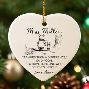 May include: Heart-shaped ceramic ornament with a gold string, featuring a Winnie the Pooh illustration and the text "Miss Miller." The ornament includes a quote and a name, perfect for a teacher gift. Christmas tree and ornaments in the background.