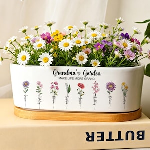 Könnte beinhalten: Ein weißer, rechteckiger Keramik-Pflanzkübel mit Bambussockel, gefüllt mit bunten Gänseblümchen und kleinen Wildblumen. Die Vorderseite zeigt den Text Grandma's Garden, Make Life More Grand sowie acht einzelne Blumenillustrationen mit Namen darunter.