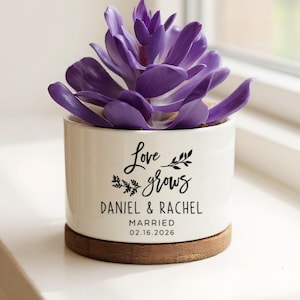 May include: A white ceramic planter with a purple succulent, sitting on a wooden base. The planter has the text "Love grows" with the names "DANIEL & RACHEL" and the date "02.16.2026" in black script. The planter is on a white windowsill.