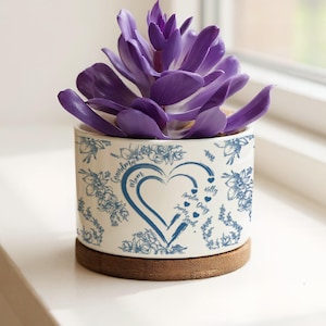 Op de afbeelding: Een witte keramische bloempot met blauwe bloempatronen en een hartvormig ontwerp, met een paarse vetplant. De pot staat op een houten onderzetter. Tekst bevat "Grandma", "Mom" en namen.