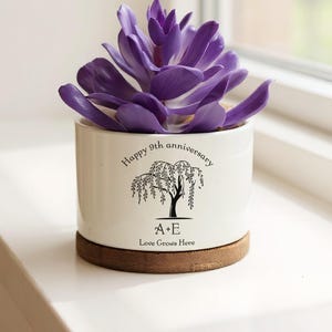 May include: A white ceramic planter with a purple succulent. The planter has a wooden base and features a black illustration of a tree with the text "Happy 9th anniversary" and "Love Grows Here".