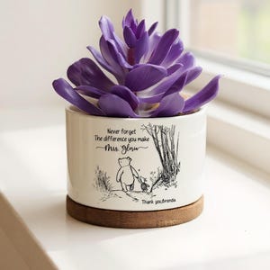 Peut inclure: Un pot en céramique blanc avec une plante succulente violette. Le pot présente une illustration de Winnie l'Ourson et le texte "Never forget the difference you make Mrs. Glow". Il repose sur un dessous de verre en bois.