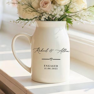 Op de afbeelding: Een witte keramische kan met een handvat, gevuld met een boeket witte en roze rozen en groen. De kan heeft de namen "Raheel & Allen", een lijn met een hart en de tekst "ENGAGED 11.06.2025". Het staat op een vensterbank.