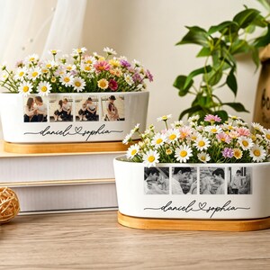 May include: Two white oval ceramic planters with bamboo bases, each filled with small white, pink, and yellow daisies. The front of each planter features four black and white printed photographs and the cursive text "daniel & sophia".