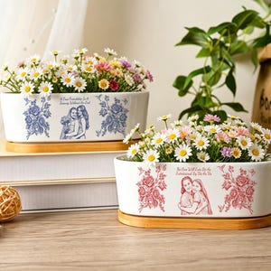 May include: Two oval white ceramic planters with bamboo bases, each filled with small white and pink daisies. One planter features blue floral patterns and text, while the other displays red floral patterns and text depicting two people hugging.
