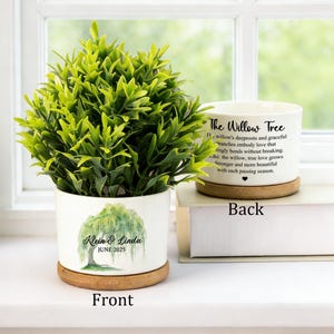 Puede incluir: Una maceta de cerámica blanca con una planta verde, con un diseño de sauce y los nombres "Klein & Linda" con "JUNIO 2025". La maceta se encuentra sobre un posavasos de madera. La parte posterior tiene texto.