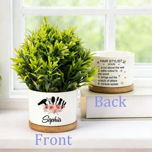 Puede incluir: Maceta de cerámica blanca con una planta verde, con un diseño floral y el nombre "Sophia". La maceta incluye un posavasos de madera. La maceta trasera tiene el texto "HAIR STYLIST" y una lista de atributos.