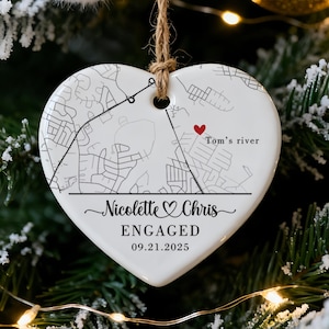 May include: Heart-shaped ceramic ornament with a map design, the words "Nicolette & Chris ENGAGED 09.21.2025" and a red heart. The ornament hangs from a twine string on a Christmas tree with other ornaments.