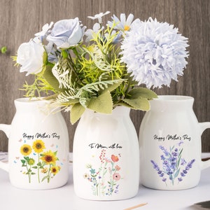 Puede incluir: Tres jarrones de cerámica blanca con diseños florales y el texto "Happy Mother's Day" y "To Mom, with love." Los jarrones contienen flores artificiales en tonos azules, blancos y verdes. Un jarrón presenta girasoles, otro flores silvestres y el último lavanda.