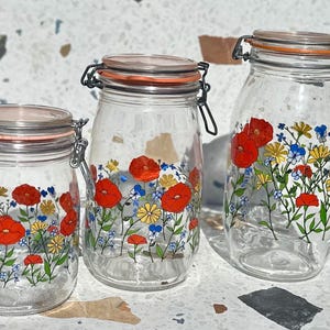 May include: Three clear glass storage jars of varying heights, each featuring a hinged lid with an orange rubber seal and a metal wire clamp. The exterior of each jar is decorated with a colorful floral pattern of red poppies, yellow daisies, and small blue flowers.