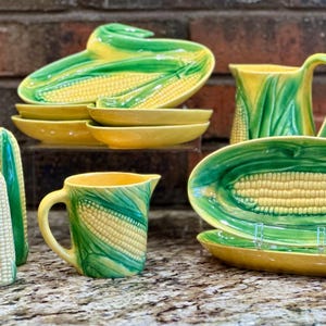 May include: A collection of ceramic corn-themed dinnerware featuring yellow corn kernels and green husks. The set includes three salt and pepper shakers, two small pitchers, several oval plates, and small bowls with embossed corn cob designs.