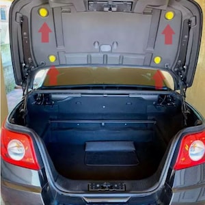 May include: A black convertible car with the trunk open. The trunk is empty and has a black mat on the floor. There are two yellow plastic caps on the trunk lid.