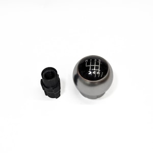 May include: A silver-colored, spherical gear shift knob with a black top displaying a six-speed shift pattern. A separate black plastic component is positioned to the left. The items are set against a white background.