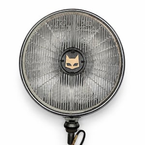 Pode incluir: Um farol automotivo redondo com aro cromado e lente de vidro estriado. O centro apresenta um emblema circular preto com um ícone de rosto de gato dourado. O texto gravado no vidro inclui 720, 700, CIBIE e Stanley. Um parafuso de montagem metálico projeta-se da parte inferior.