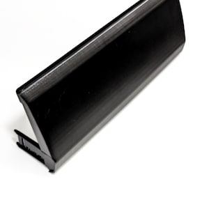 Puede incluir: Un objeto rectangular negro con una superficie texturizada y un pequeño accesorio en ángulo. El objeto parece estar hecho de un material sólido y está sobre un fondo blanco. El diseño es elegante y moderno.