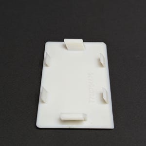 Può includere: Un componente in plastica rettangolare bianco con quattro linguette sporgenti e una linguetta rettangolare. Il numero "7700764476" è stampato verticalmente sul lato destro del componente.
