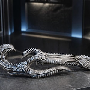 Puede incluir: Un arma de utilería plateada con un diseño biomecánico intrincado. Presenta elementos curvos en forma de garra y una superficie texturizada, lo que sugiere una estética futurista o alienígena. El arma se exhibe sobre una superficie oscura.
