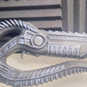 Könnte beinhalten: Eine detaillierte, graue Requisitenwaffe mit einem biomechanischen Design. Die Requisite verfügt über einen gebogenen, strukturierten Griff und eine Reihe scharfer, klingenartiger Vorsprünge entlang ihrer Länge. Die Gesamtästhetik deutet auf ein Science-Fiction- oder Fantasy-Thema hin.