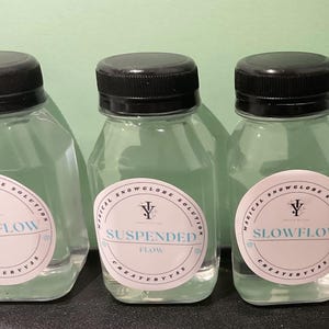 Puede incluir: Tres botellas de vidrio transparente con tapas negras y etiquetas blancas. Las etiquetas dicen "Magical Snowglobe Solution" y tienen diferentes velocidades de flujo: "Fast Flow", "Suspended Flow" y "Slow Flow".