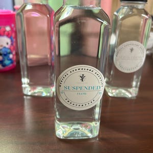 Puede incluir: Botellas de plástico transparente con tapas negras, llenas de un líquido transparente. Las botellas tienen etiquetas blancas con el texto "Magical Snowglobe Solution" y "Suspended Flow". Las botellas están sobre una superficie marrón.