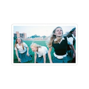 May include: A sticker featuring a photograph of four young women in school uniforms running on a green field. They are wearing white collared shirts, dark blue skirts, and some have black vests. The background includes a building and a light blue sky.