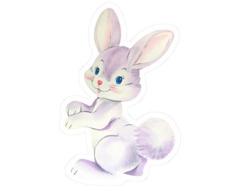 May include: A watercolor illustration of a cute bunny rabbit in a seated pose. The bunny has lavender fur, large ears, blue eyes, and a fluffy white tail. The rabbit is smiling with a pink nose and tongue, and is set against a white background.