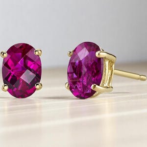 Op de afbeelding: Een paar gouden oorstekers met ovaal geslepen, diep magenta edelstenen. De edelstenen zijn gezet in een vier-klauwzetting en de oorbellen hebben een klassieke oorbel met een vlindersluiting.
