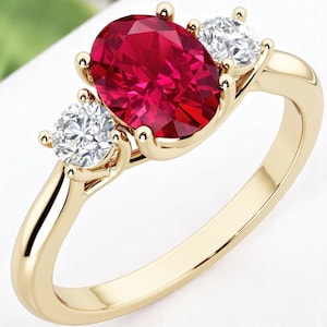 Op de afbeelding: Een gouden ring met een grote ovale robijn als centrale steen, geflankeerd door twee kleinere, ronde, heldere edelstenen. De ring heeft een klassiek ontwerp met drie stenen en een gepolijste gouden band.