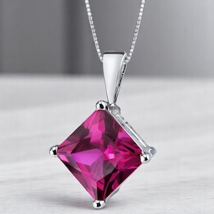 May include: A silver pendant necklace featuring a large, square-cut, vibrant magenta gemstone held by four prongs. The pendant is suspended from a delicate silver chain, showcasing a faceted surface that reflects light across the stone's deep pink hue.