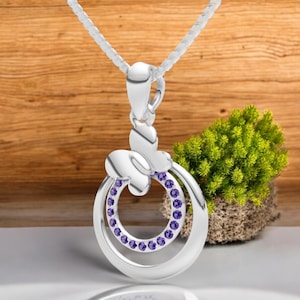 May include: A silver pendant necklace featuring a butterfly design. The pendant has a double-circle design with purple gemstones and a butterfly accent. The necklace hangs on a white chain, with a wooden background.