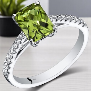Può includere: Un anello d'argento con una grande pietra di peridoto verde chiaro rettangolare. La fascia è adornata da piccole pietre preziose trasparenti. L'anello è presentato su uno sfondo sfocato, evidenziando i dettagli del gioiello. L'anello è contrassegnato con "14k".