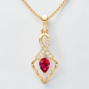 Op de afbeelding: Een gouden hangerketting met een druppelvormige, dieprode edelsteen. De hanger heeft een decoratief open ontwerp met kleine, heldere edelstenen en hangt aan een fijne gouden ketting. De ketting staat tegen een witte achtergrond.