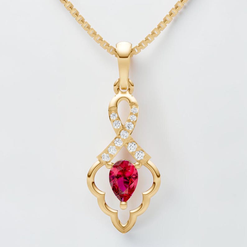 May include: A gold pendant necklace featuring a teardrop-shaped, deep red gemstone. The pendant has a decorative openwork design with small, clear gemstones and is suspended from a delicate gold chain. The necklace is set against a white background.