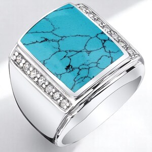 May include: A silver ring with a rectangular turquoise stone center. The turquoise stone has dark veining. Small, clear stones are set along the top and bottom edges of the turquoise stone. The ring has a polished, shiny finish.