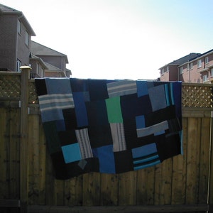 May include: A blue and green patchwork quilt hanging on a wooden fence. The quilt is made of different shades of blue and green fabric, and it has a simple, geometric design.