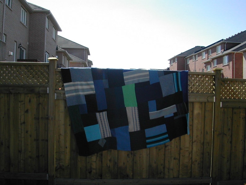 May include: A blue and green patchwork quilt hanging on a wooden fence. The quilt is made of different shades of blue and green fabric, and it has a simple, geometric design.
