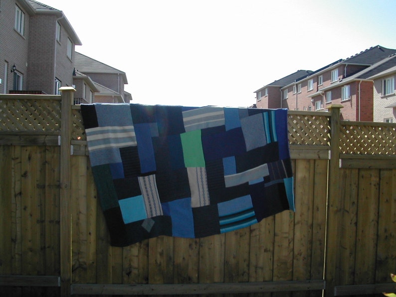 May include: A blue and green patchwork quilt hanging on a wooden fence. The quilt is made of different shades of blue and green fabric, and it has a variety of patterns.