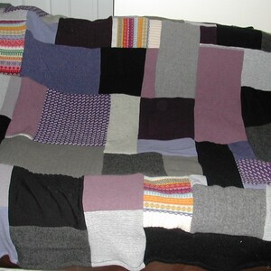 May include: A patchwork quilt made from different colored knitted fabrics. The quilt is made up of squares and rectangles of various sizes and colors, including gray, purple, black, and white.